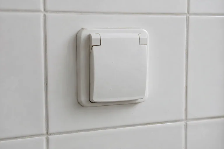 Wit stopcontact met gesloten beschermklep op een betegelde wand.