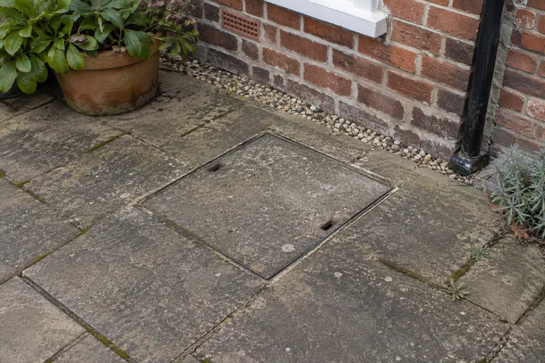 Gesloten inspectieluik in de bestrating naast een gevel.