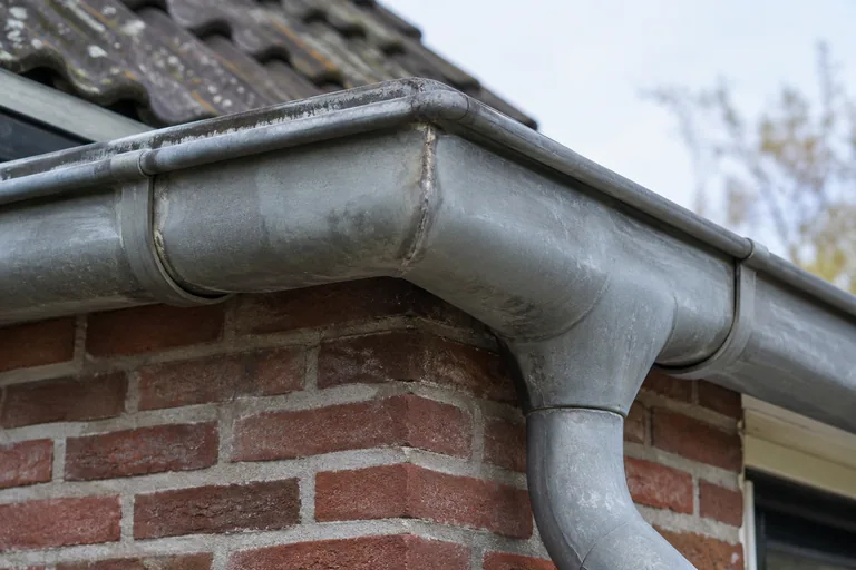 Verweerde zinken dakgoot met een hoek en regenpijpaansluiting.