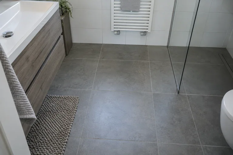 Grijze tegelvloer in een badkamer met douche en wastafelmeubel.